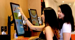 Two smiling women in a busy restaurant place an order on a self-service touchscreen kiosk
